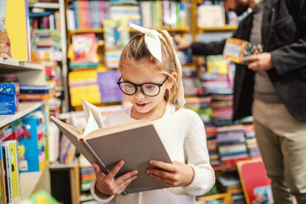 Young girl wearing glasses and a white hair bow reads a large-print book in a library surrounded by colorful shelves of books.