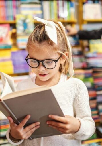 Young girl wearing glasses and a white hair bow reads a large-print book in a library surrounded by colorful shelves of books.